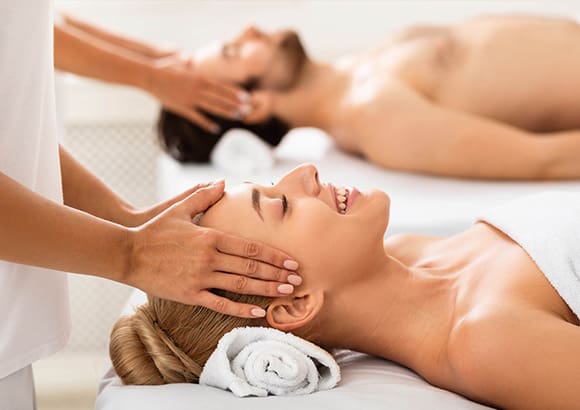 Couple lying face up on two massage tables, draped in white sheets, receiving head massages from two therapists