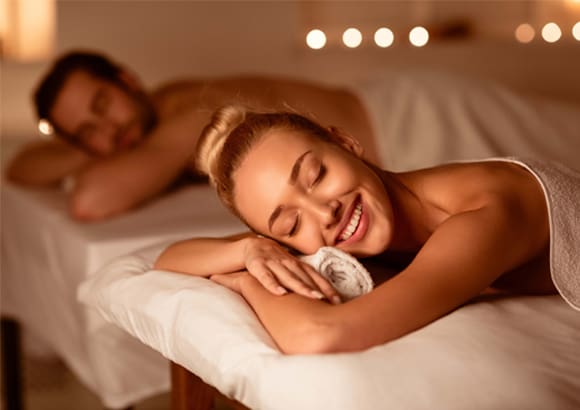 Couple lying side by side on two massage tables, draped in white sheets, in a dimly lit room