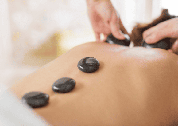 Woman lying face down, draped in white sheets, warm stones along her back, therapist massaging her trapezius with stones