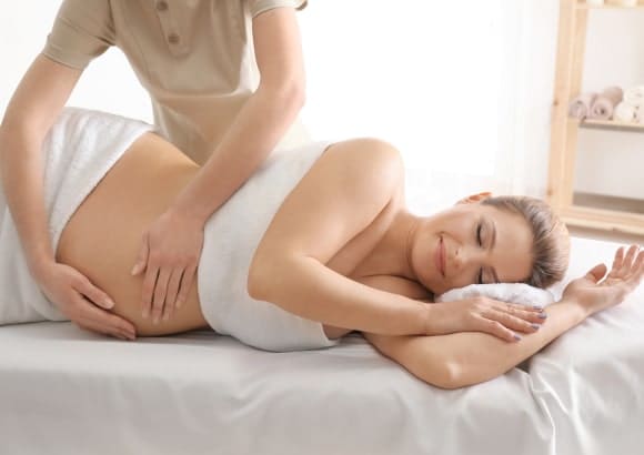 Pregnant woman in side-lying position draped in a white sheet, therapist gently massaging her belly